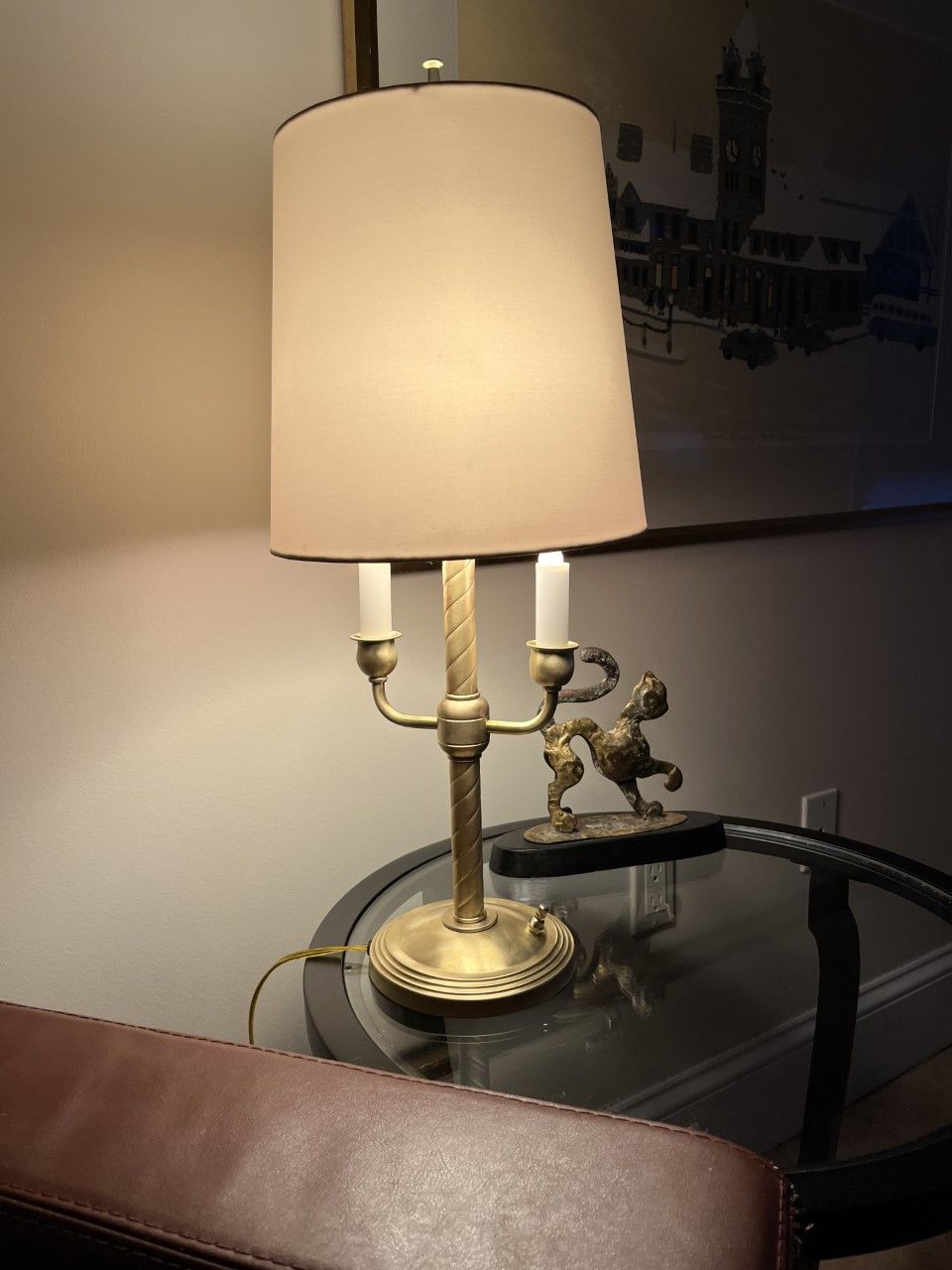 Gold lamp with a tan shade, on a glass table. A statue of a horse is on the table beside it.