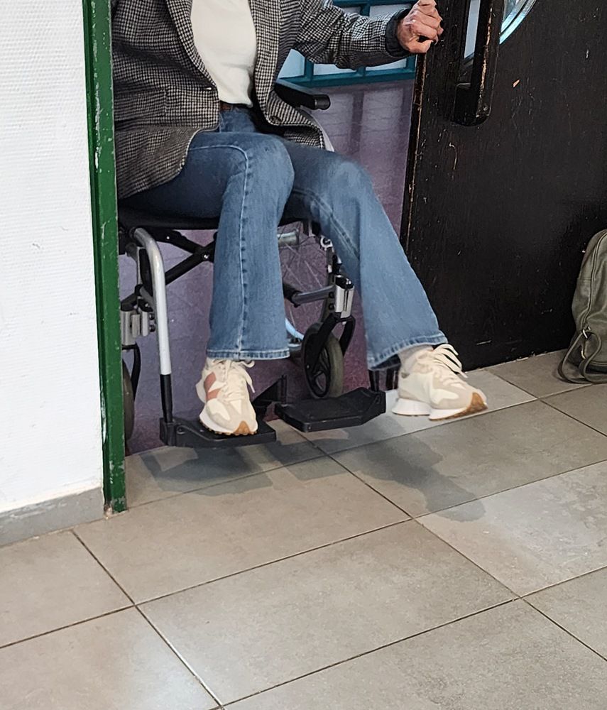 אדם יושב בכיסא בתוך הבית, לובש ג'ינס כחול ונעלי ספורט לבנות, ליד קיר שחור ורצפה מרוצפת