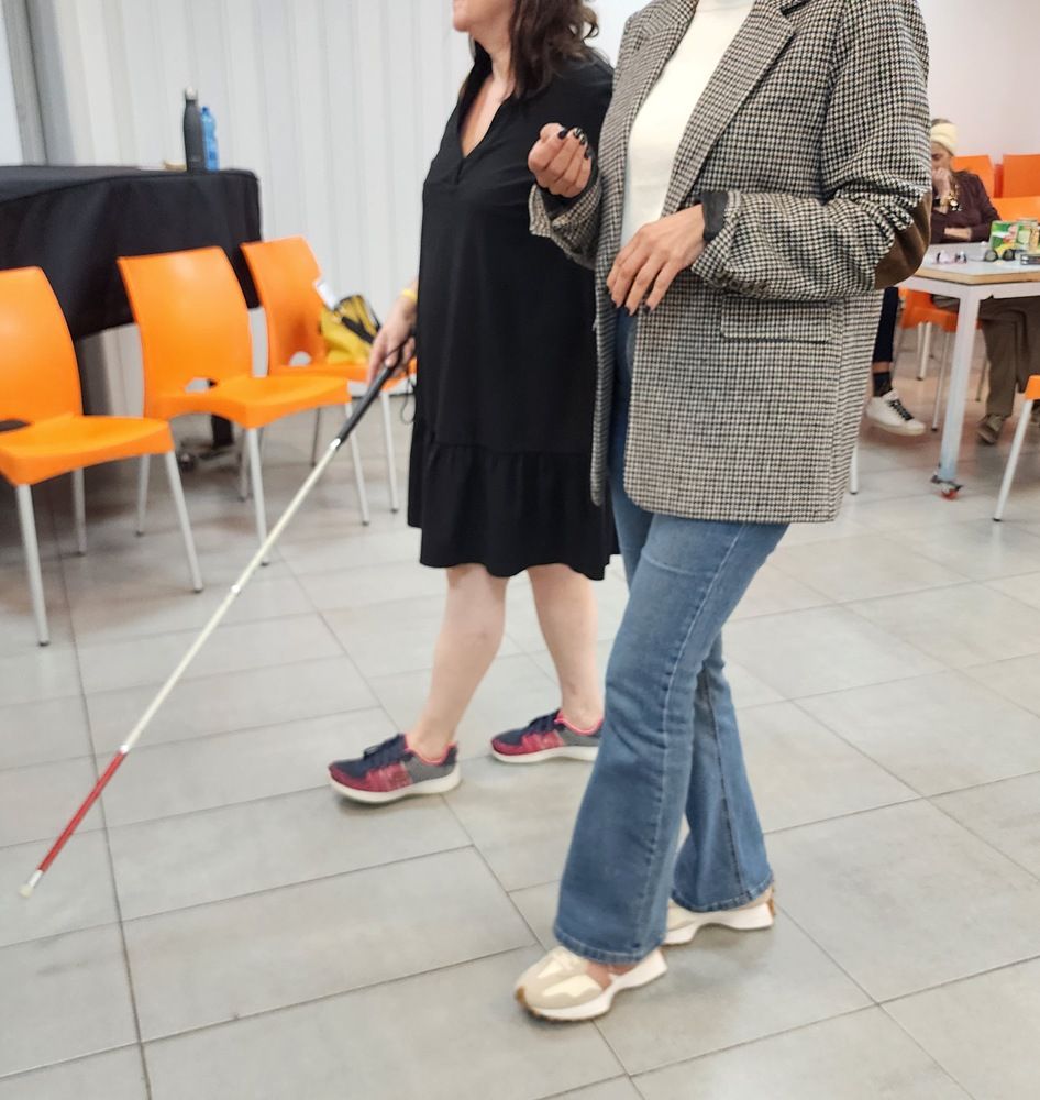שתי נשים הולכות בתוך הבית, אחת משתמשת במקל הליכה לבן, בחדר עם כיסאות כתומים.