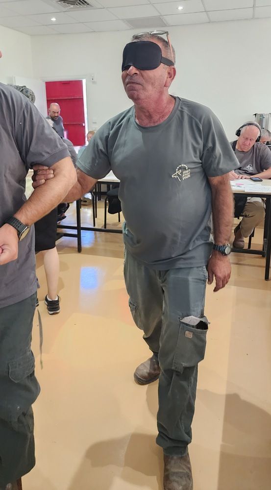 גבר עטור עיניים בבגדים אפורים הולך בחדר מואר עם אנשים יושבים סביב שולחנות