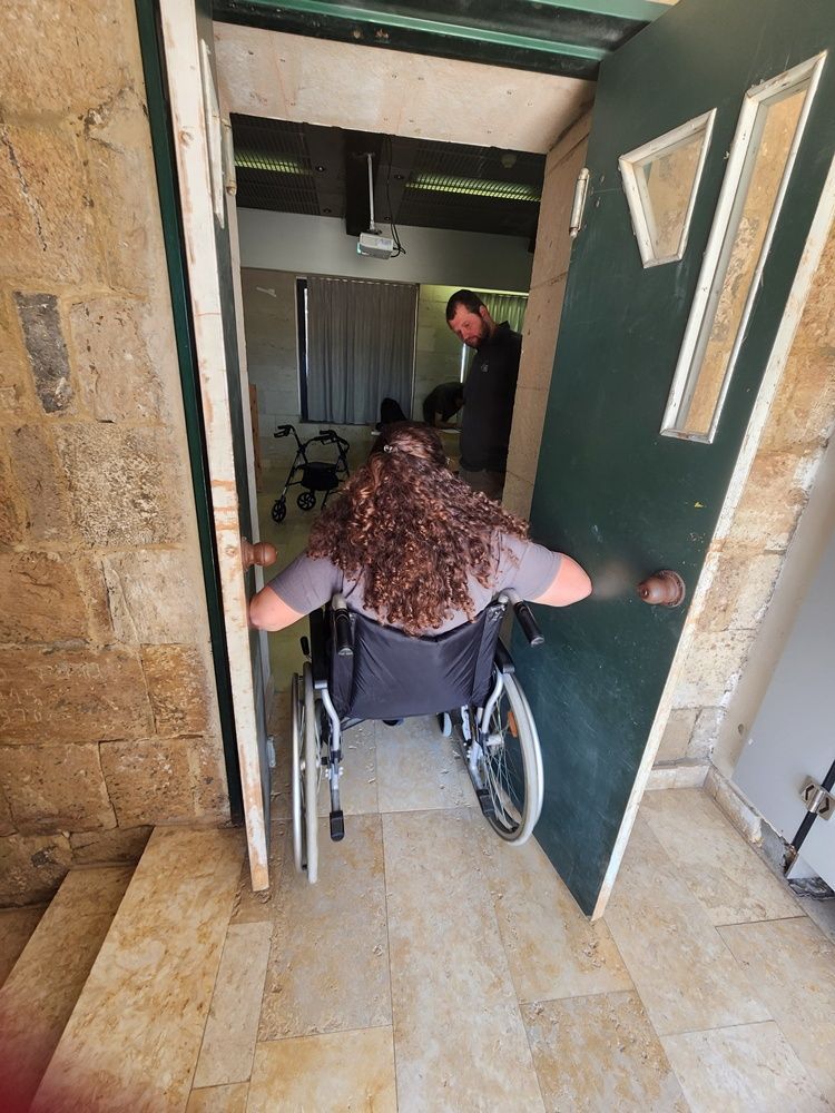 אדם בכיסא גלגלים נכנס דרך פתח צר, כשאדם נוסף לפניו במסדרון.