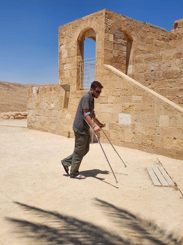 אדם המשתמש בקביים הולך ליד בניין אבן מואר שטוף שמש עם פתח מקומר.