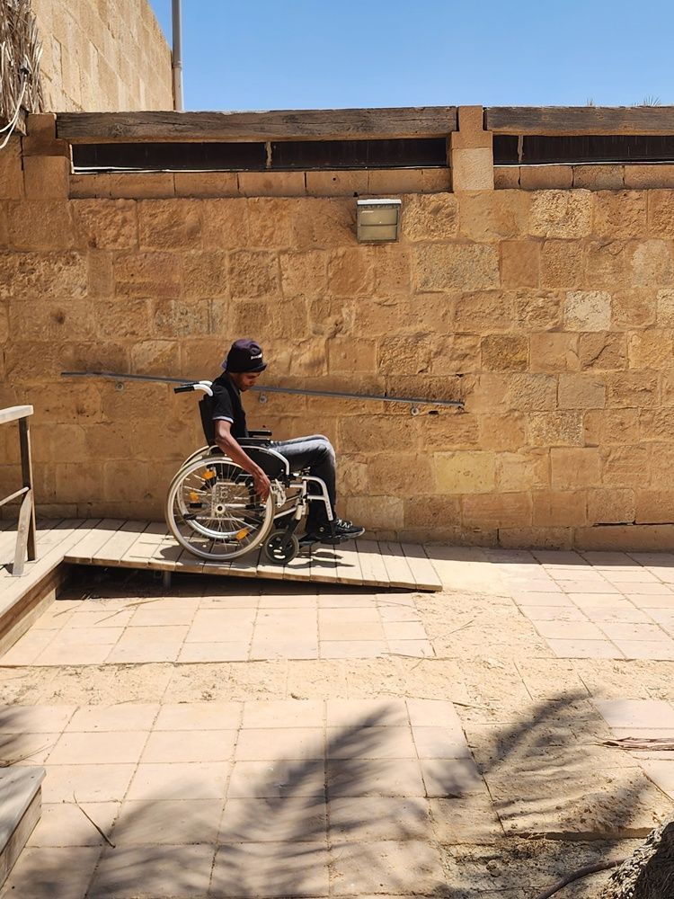 אדם בכיסא גלגלים בחוץ ליד קיר אבן, על רמפה באור שמש בהיר.