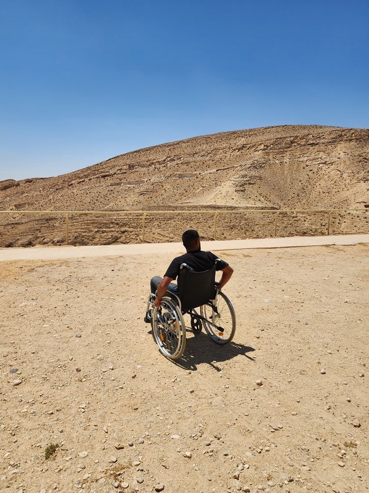 אדם בכיסא גלגלים על שביל מדברי חולי מול גבעה סלעית תחת שמיים כחולים בהירים