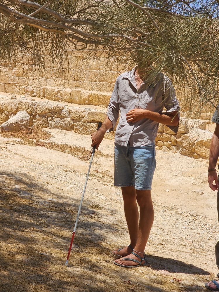 אדם עומד על שביל סלעי עם מקל הליכה מתחת לעץ, לובש חולצה אפורה ומכנסי ג'ינס קצרים.
