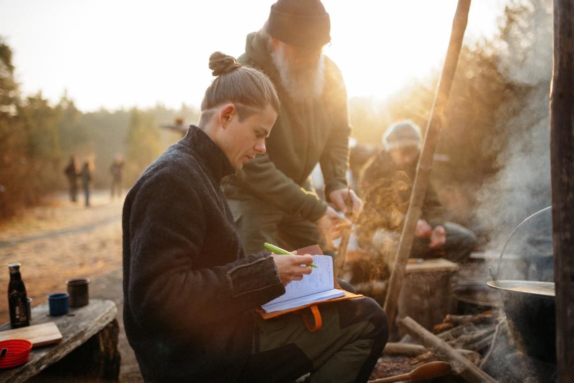 Eine Person kniet in einem Wald und sägt mit einer langen Handsäge einen von einem Holzrahmen gestützten Baumstamm durch.