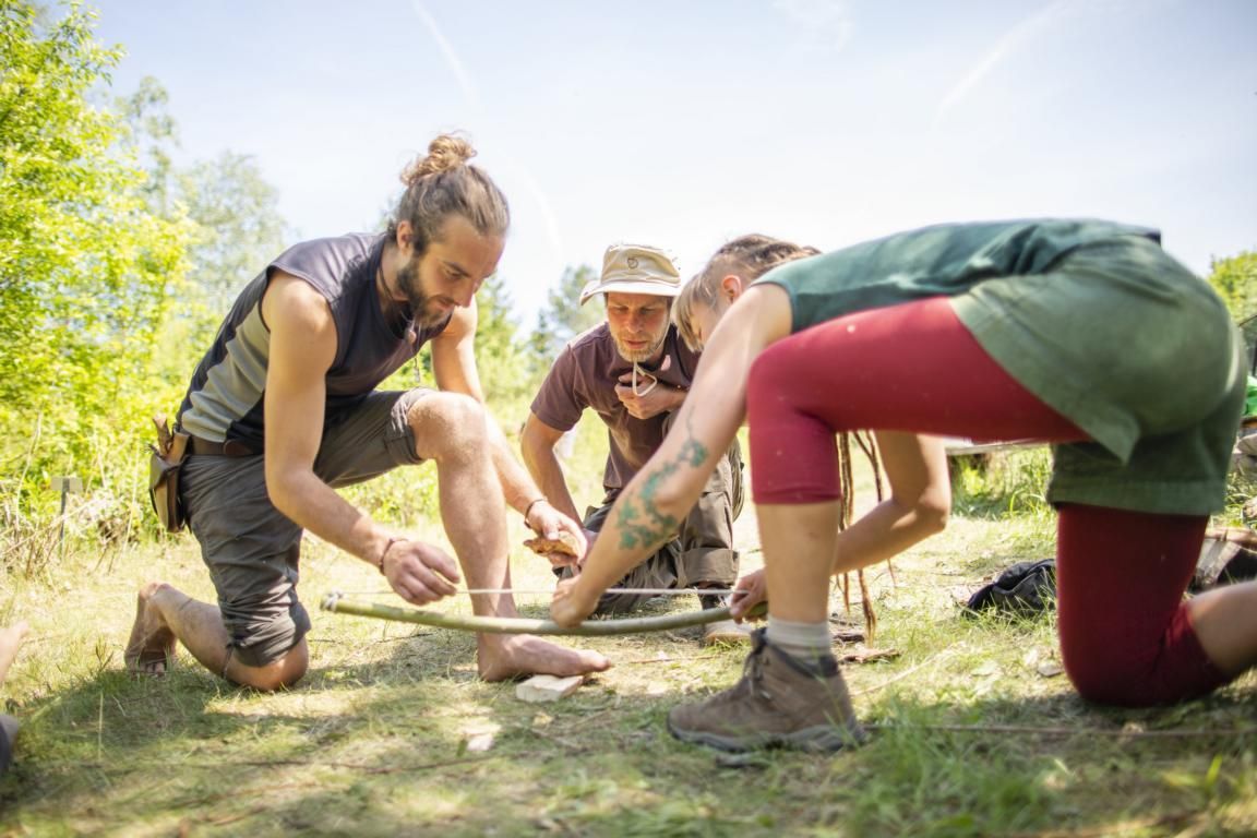 Eine Gruppe von Leuten kniet im Gras beim Feuerbohren