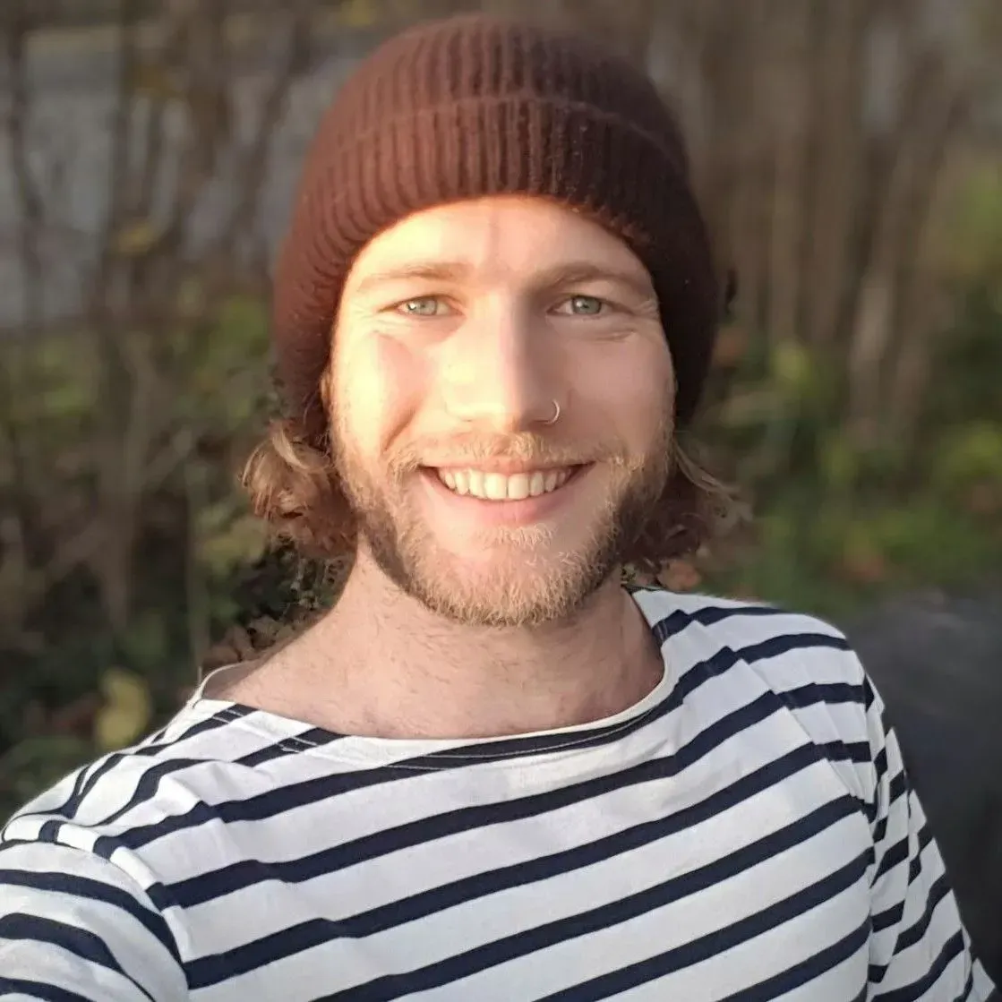 Une personne souriante, coiffée d'un bonnet marron et vêtue d'une chemise à rayures, se trouve à l'extérieur, baignée par une douce lumière du soleil.