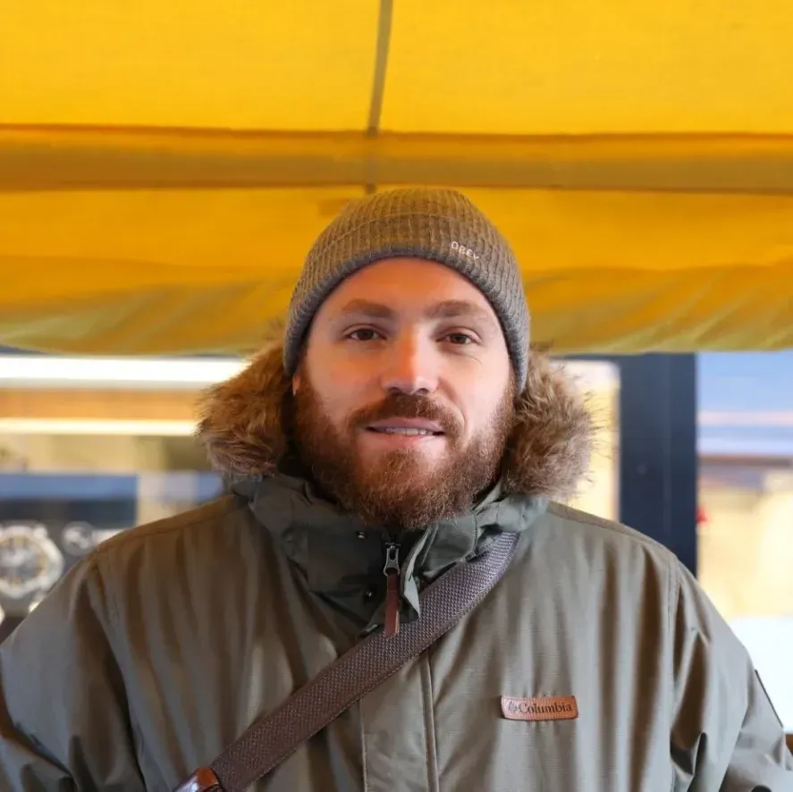 Une personne portant un bonnet et une veste gris se tient sous un auvent jaune et regarde l'objectif.