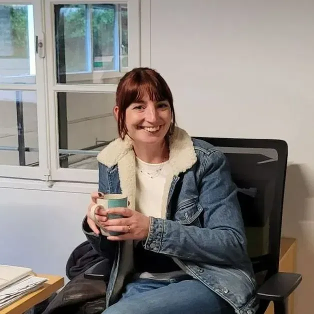 Une femme souriante, assise sur une chaise de bureau, tient une tasse près d'une fenêtre.