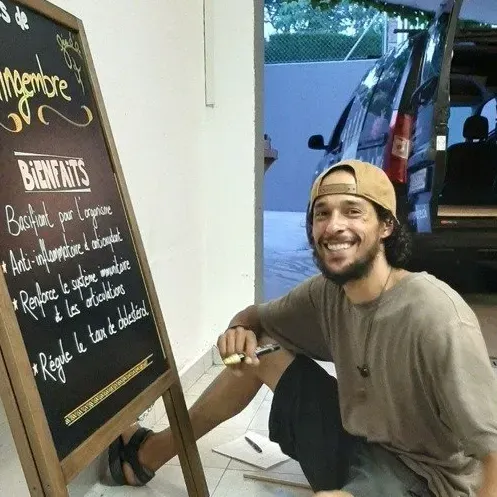 Un homme souriant, coiffé d'une casquette beige, est accroupi près d'un tableau noir devant un café.