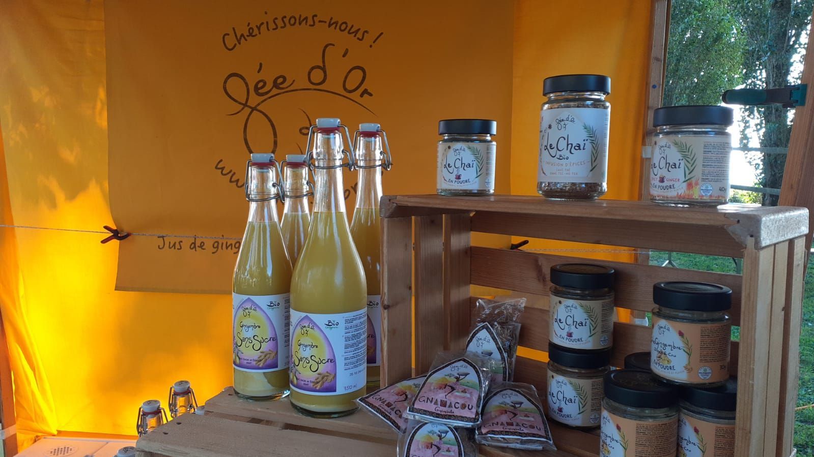 Étal de marché avec des bocaux, des bouteilles et des produits emballés sur des étagères en bois sous un auvent jaune.