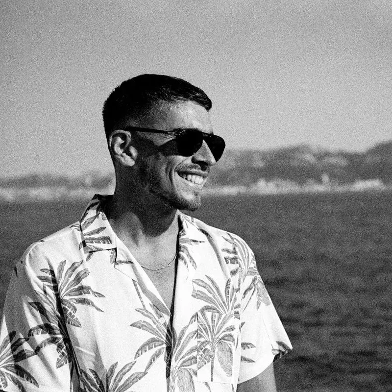 Un homme portant des lunettes de soleil et une chemise à motifs sourit au bord de l'océan, avec le rivage à l'horizon.