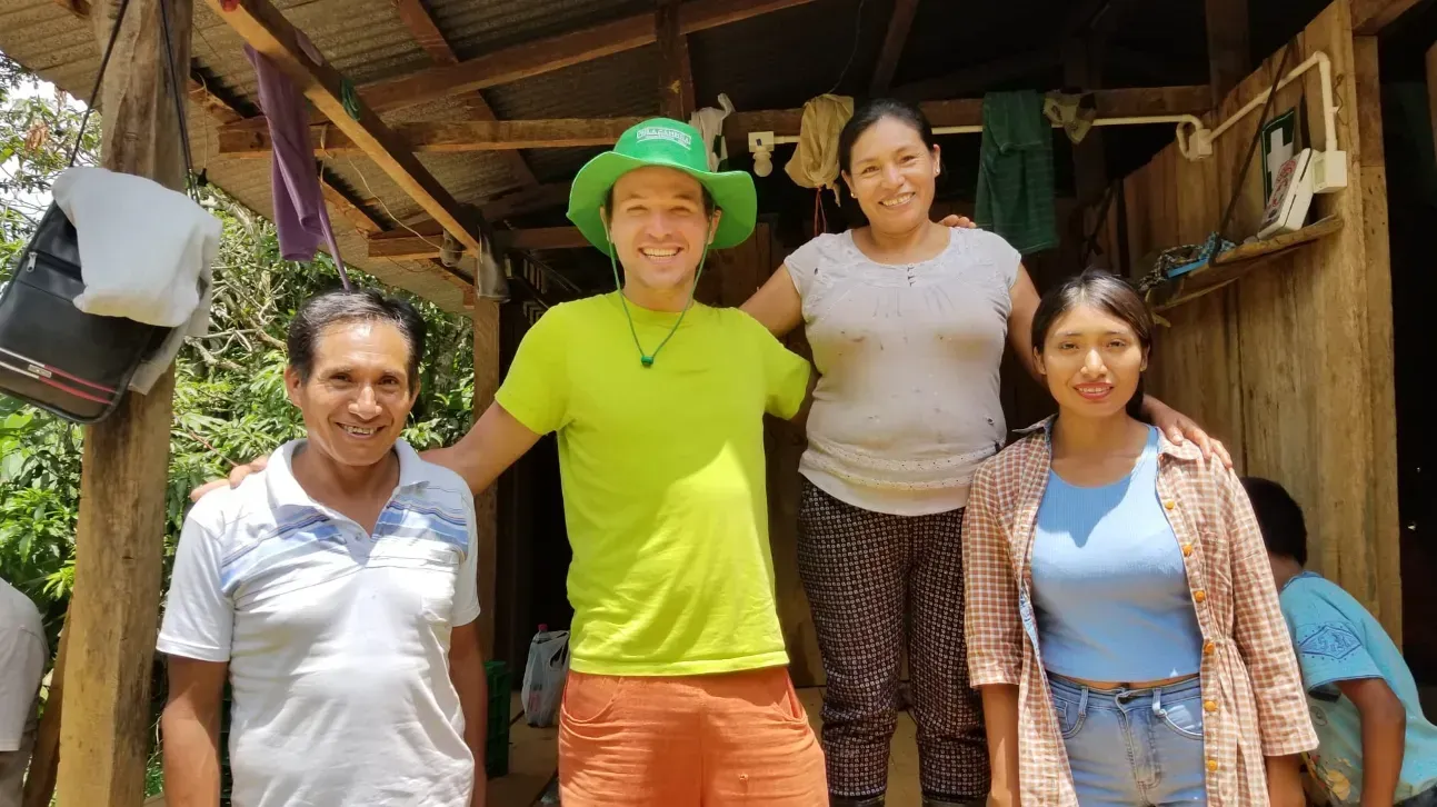 Quatre personnes sourient devant une maison rustique en bois, l'une d'elles porte un chapeau vert et une chemise aux couleurs vives.