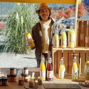 Un vendeur souriant derrière son étal de marché, entouré de bocaux et de bouteilles de boissons jaunes et de conserves, est installé à l'extérieur.