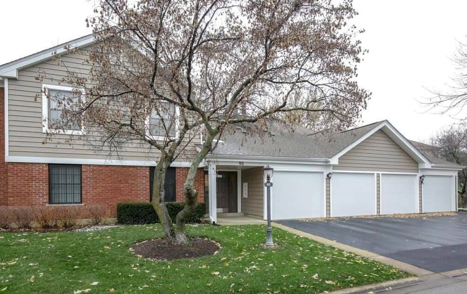 A multi-unit building featuring red brick, beige siding, a bare tree in the front lawn, and a row of three garage doors.