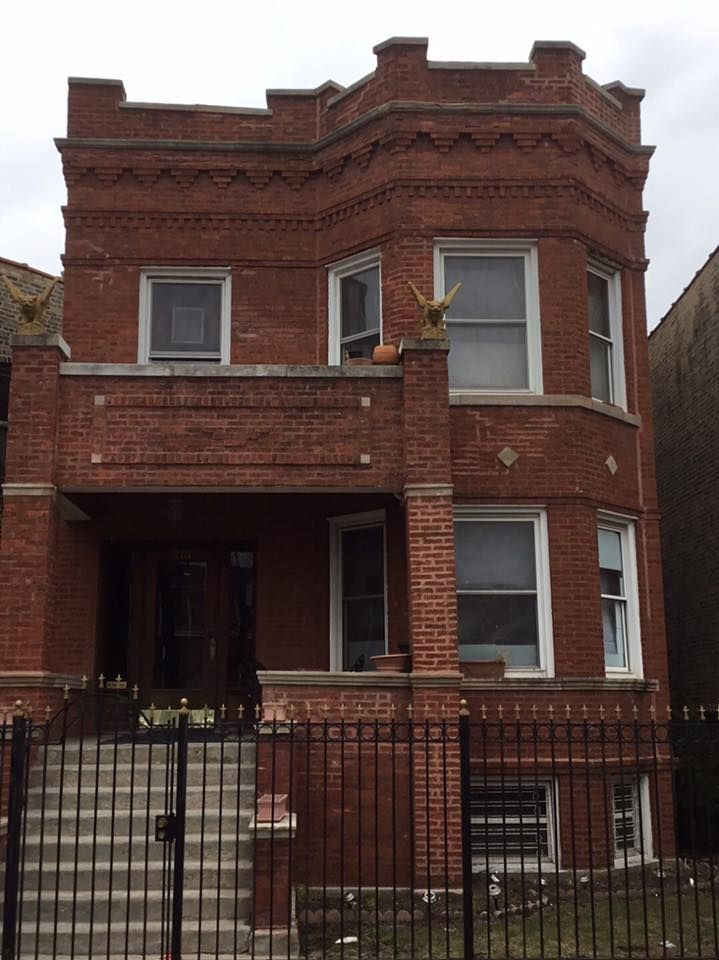 A two-story red brick house with a front porch, a bay window, a crenellated roofline, and a black metal fence.