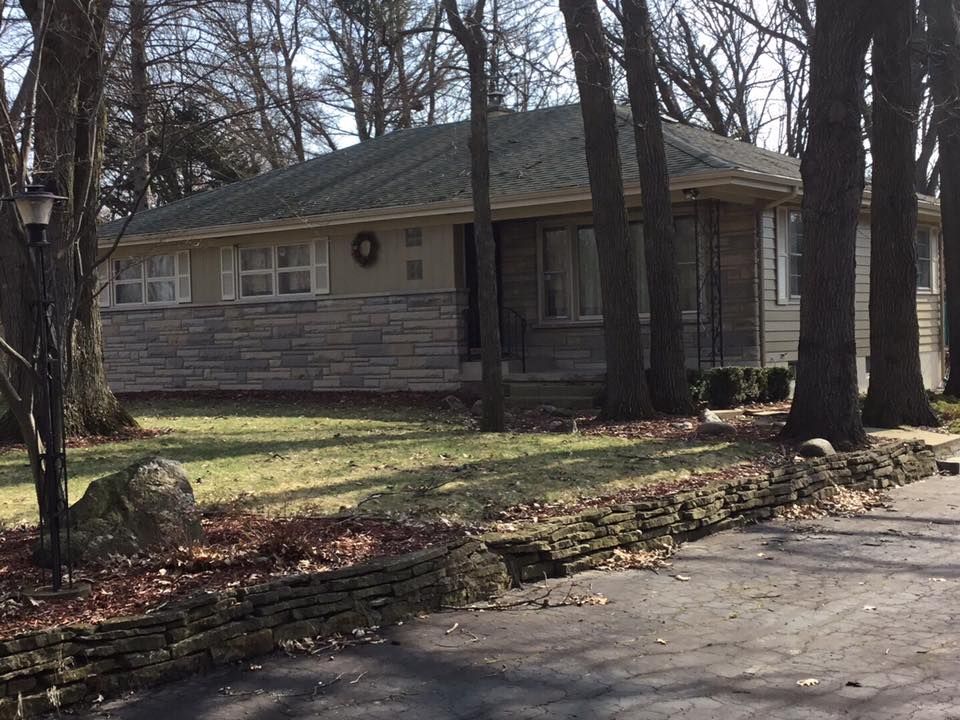 A single-story house with stone siding, a gray shingled roof, and a low stone retaining wall in front, surrounded by trees.