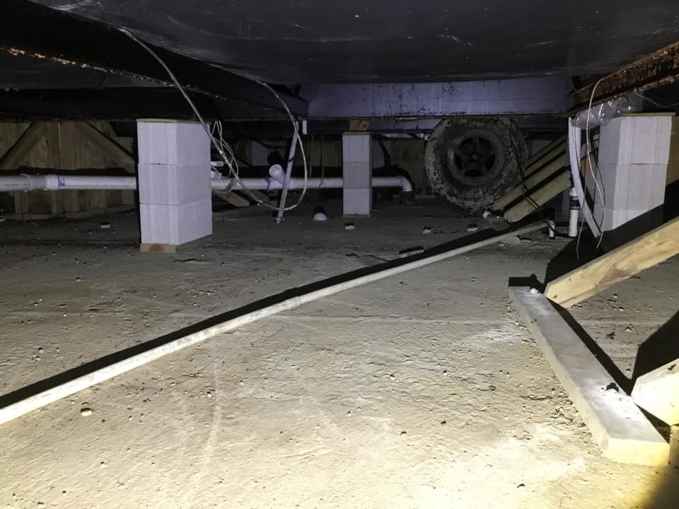The crawl space under a mobile home shows white concrete block piers supporting a dark metal chassis and PVC piping.