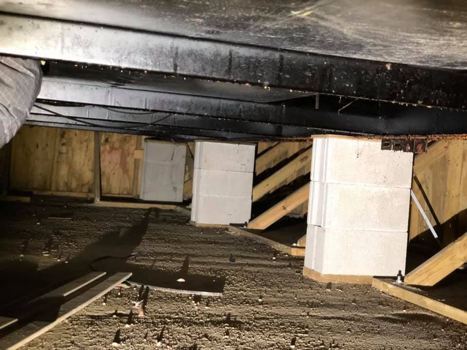 A crawl space with metal beams supported by stacks of white concrete blocks over a dark, gravel-covered ground.