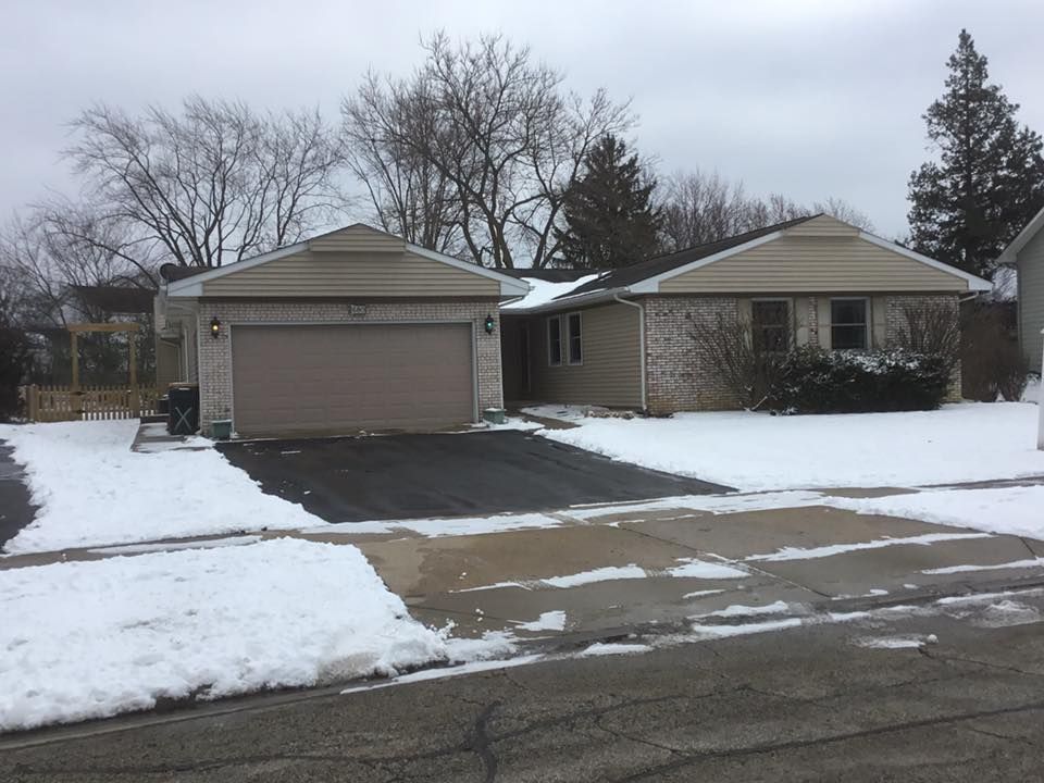 A one-story suburban home with a two-car garage, beige siding, and light brick, surrounded by snow on an overcast day.