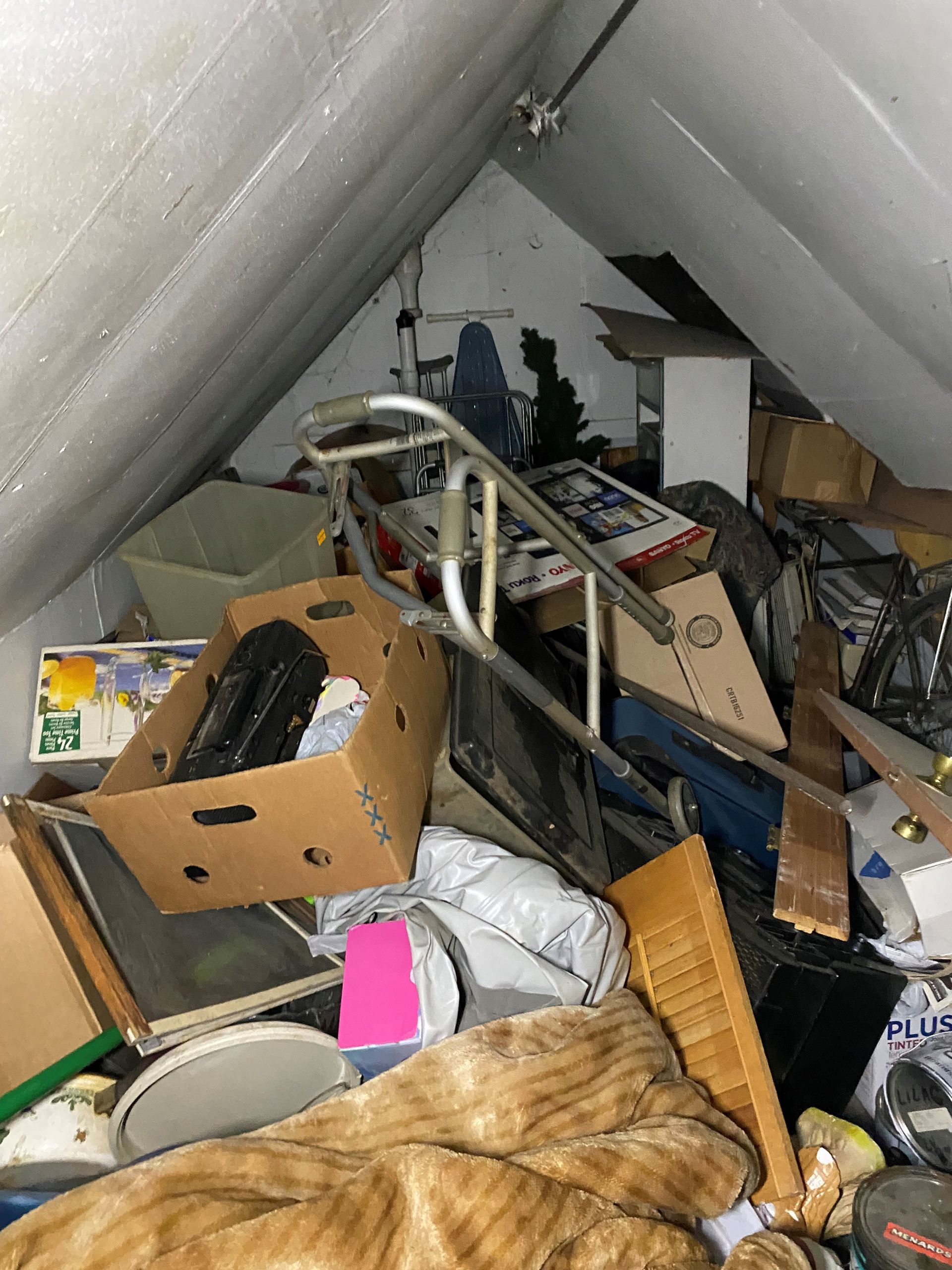 A cluttered, cramped attic storage space filled with cardboard boxes, a rolling walker, household items, and bedding.