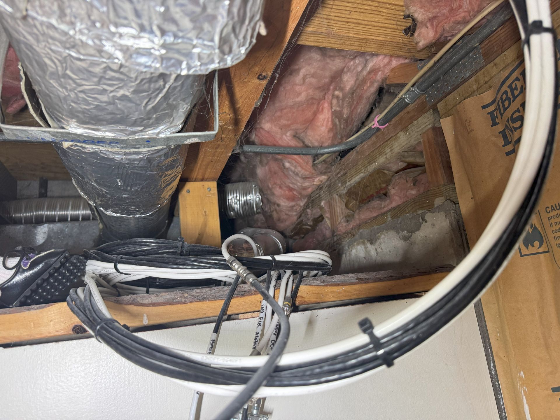 A view into an attic space with wooden joists, pink insulation, silver ductwork, and bundled black and white electrical wires.
