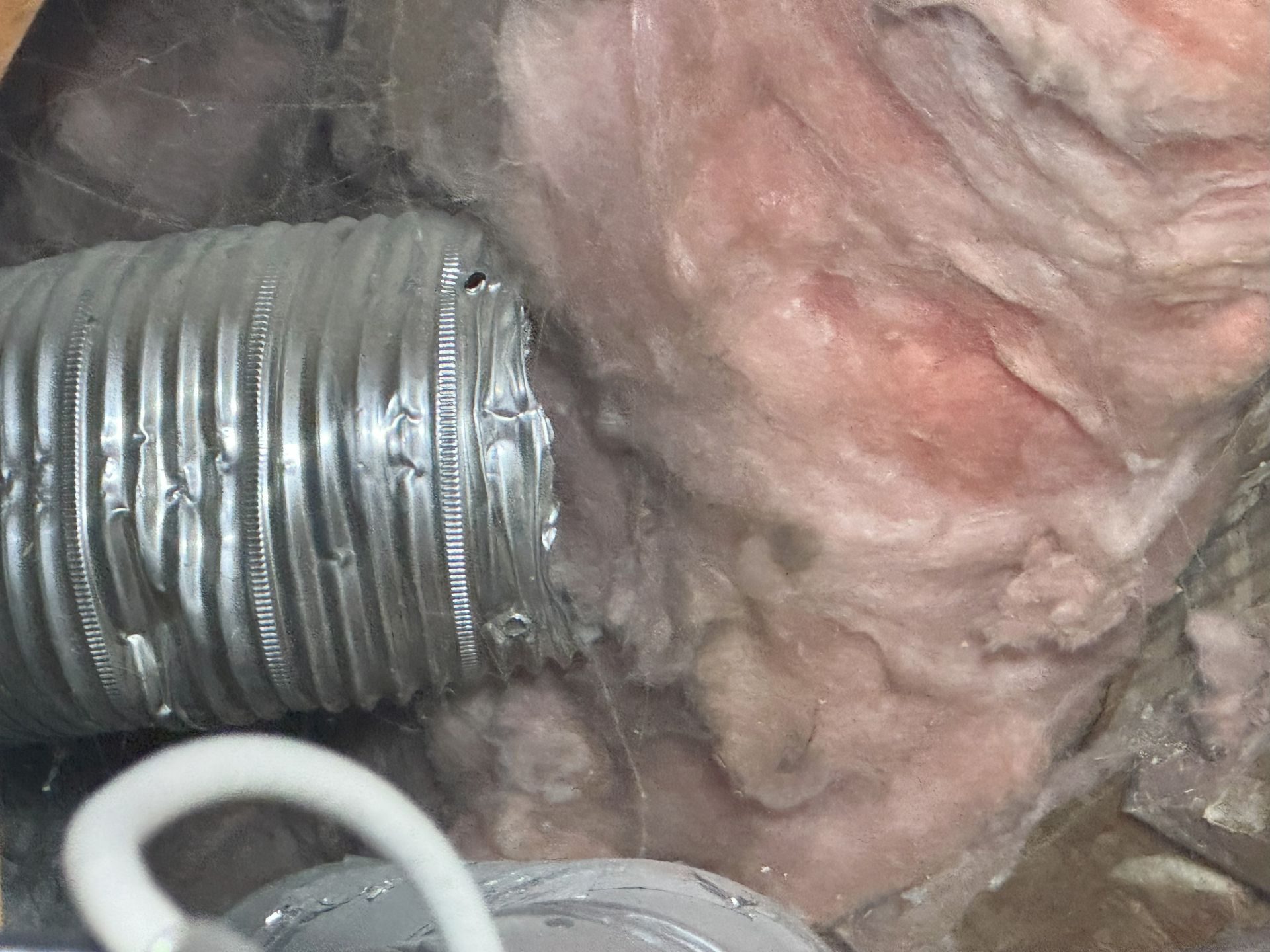 A corrugated metal duct ending abruptly next to pink fiberglass insulation inside an attic space.