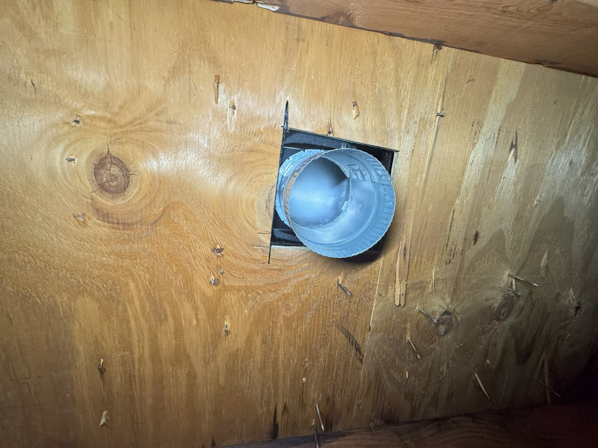 A round metal vent pipe passes through a square cutout in a plywood sheet.