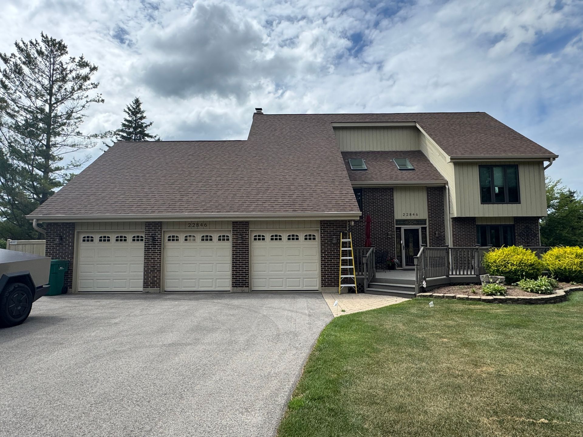 A two-story suburban home with a three-car garage, brown roof, and tan siding, set in a yard with a gravel driveway.