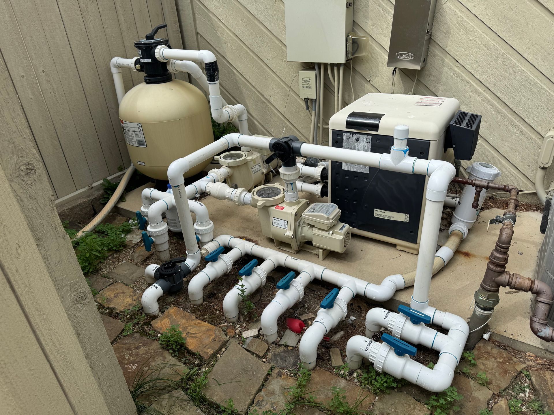 Pool equipment setup with a beige sand filter, pump, and heater connected by white PVC pipes with multiple blue valves.