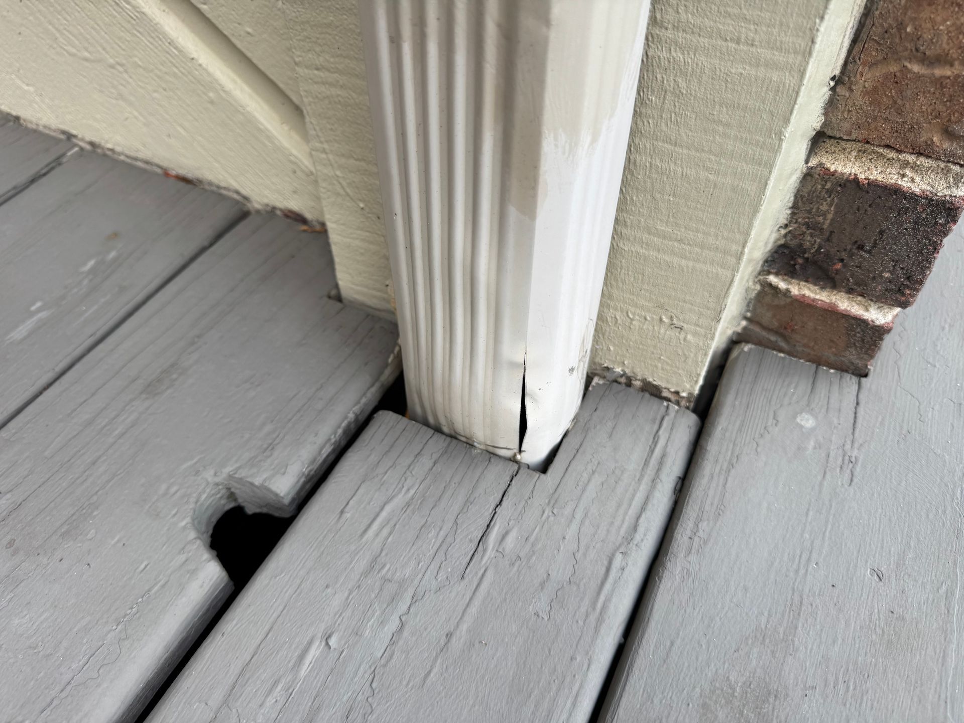 A white downspout meets gray wooden deck boards with a cutout gap, positioned against a beige wall and brick corner.