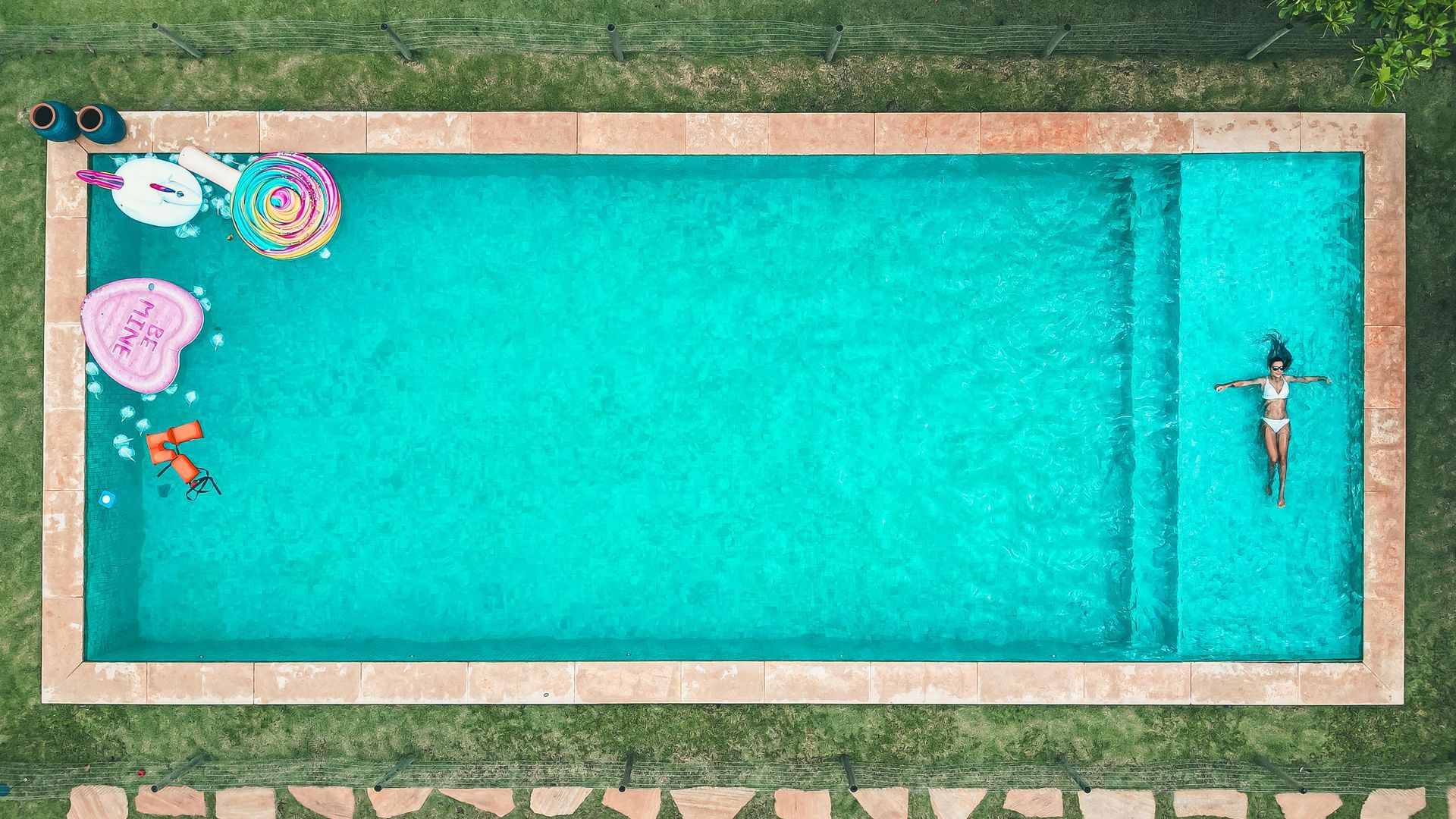 An aerial view of a person floating in a rectangular swimming pool with several colorful inflatable pool toys nearby.