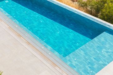 A high-angle view of a rectangular swimming pool with turquoise tiled water, next to a beige patio and green foliage.