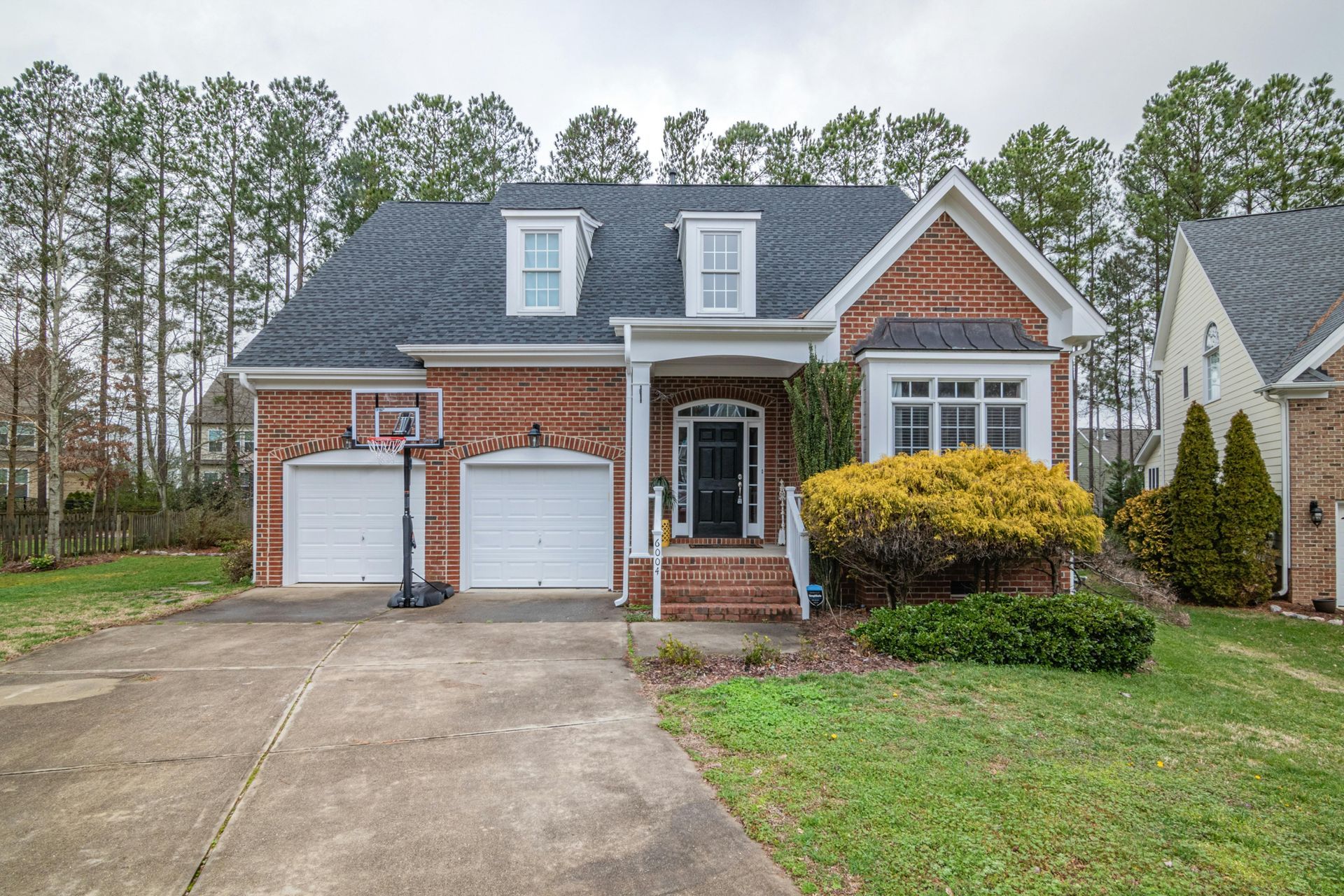 A two-story brick suburban house with a two-car garage, a concrete driveway, and dormer windows on a cloudy day.