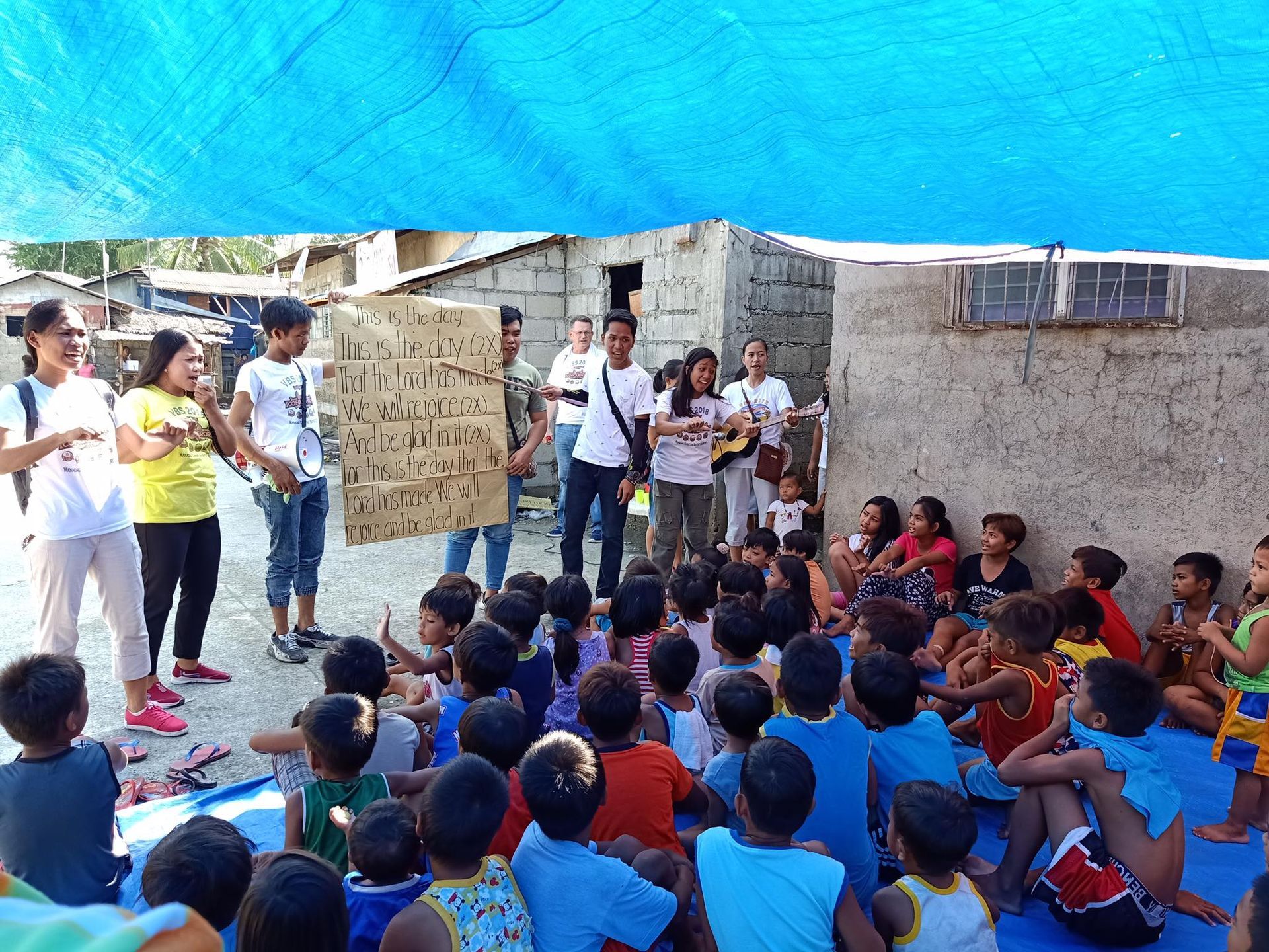 A group of children are sitting under a blue tarp