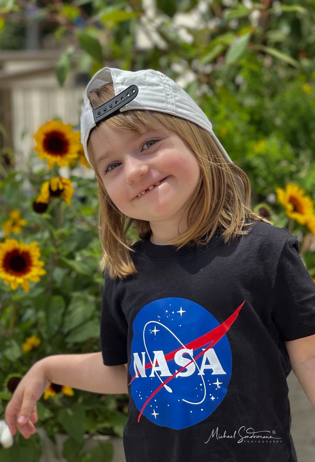 Ein kleines Mädchen mit einem NASA-T-Shirt und einem Hut steht vor Sonnenblumen.