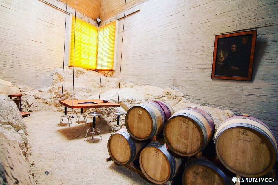 Bodega con barriles de madera apilados, mesas y ventanas iluminadas con luz amarilla en una habitación rústica.