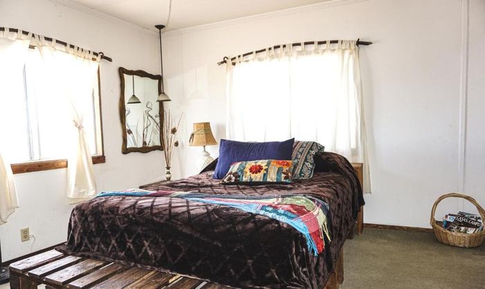 Dormitorio con una cama vestida con mantas de colores, cortinas blancas, espejo y una lámpara junto a la ventana.