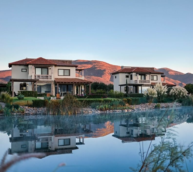 Dos casas junto a un estanque tranquilo al atardecer, con las montañas reflejadas en el agua.
