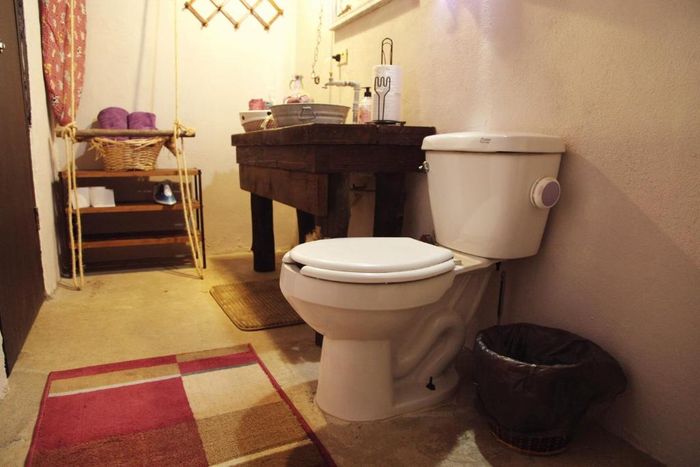 Baño con inodoro, lavabo de madera, cesta de mimbre y paredes de color beige sobre suelo de baldosas.