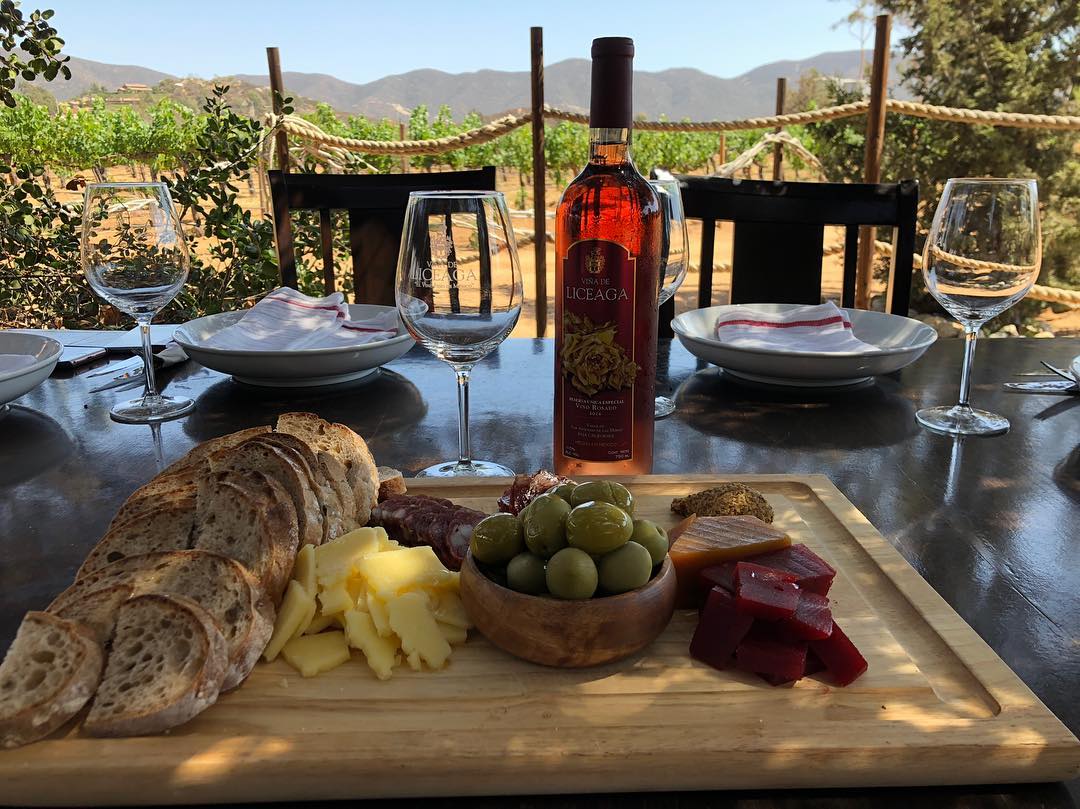 Mesa puesta con pan, fruta, botella de vino y dos juegos de cubiertos al aire libre con vistas a un viñedo y a las montañas.