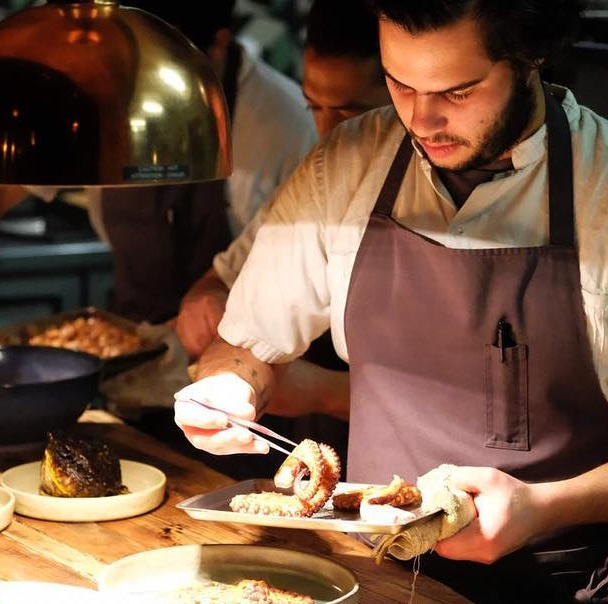 Chef emplatando comida en el mostrador de un restaurante, con delantal puesto bajo una iluminación cálida.