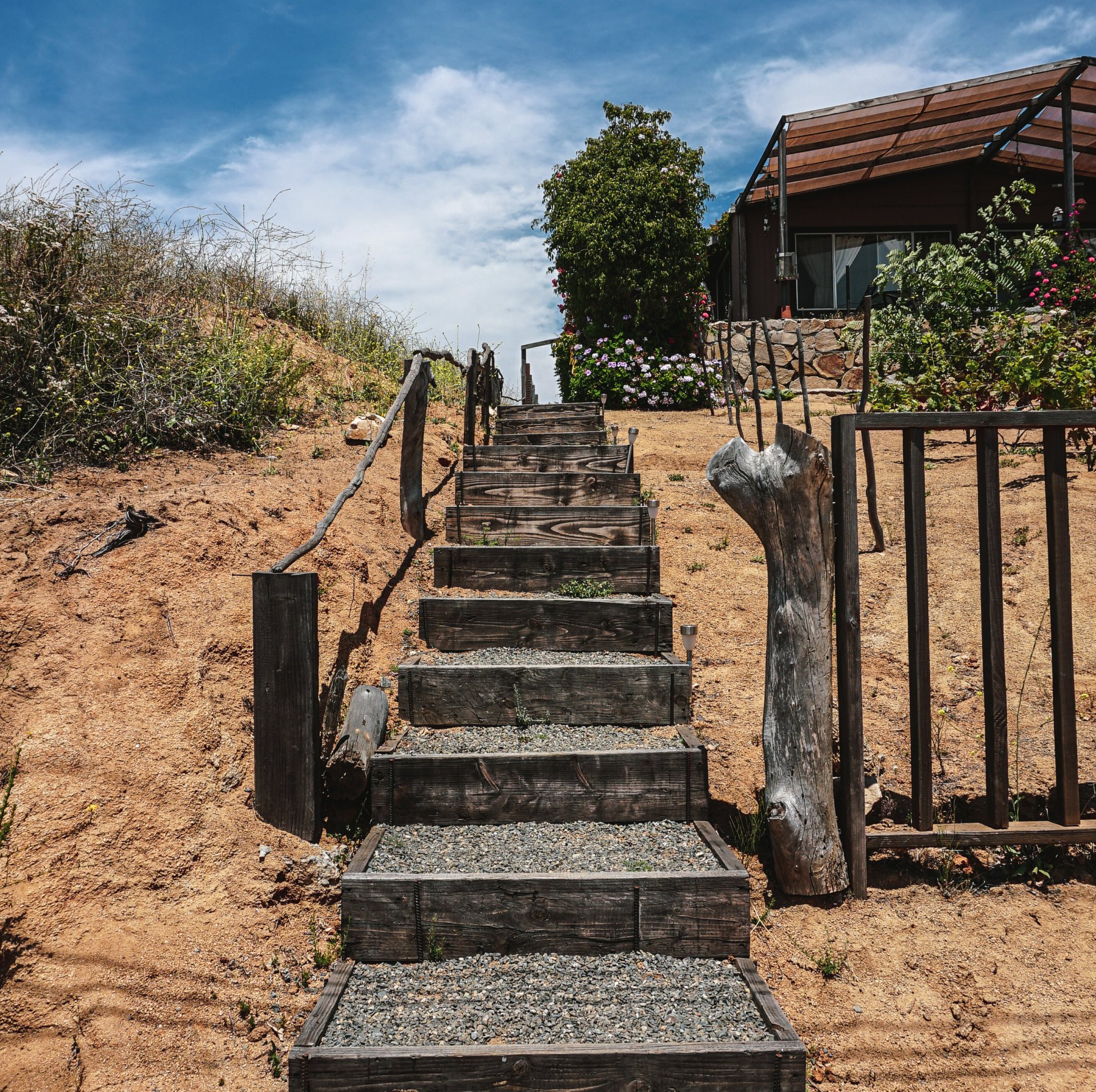 Escaleras de hormigón que suben una colina arenosa junto a una casa y una valla bajo un cielo azul.