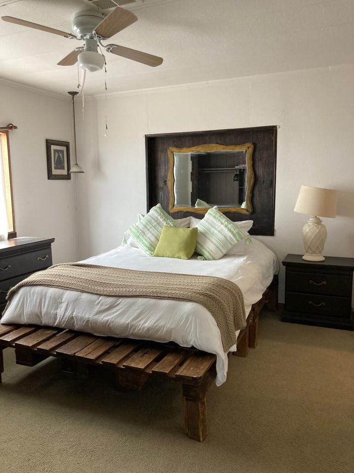 Dormitorio con cama de madera, ropa de cama blanca, almohadas, cabecero con espejo, ventilador de techo y lámparas de noche.