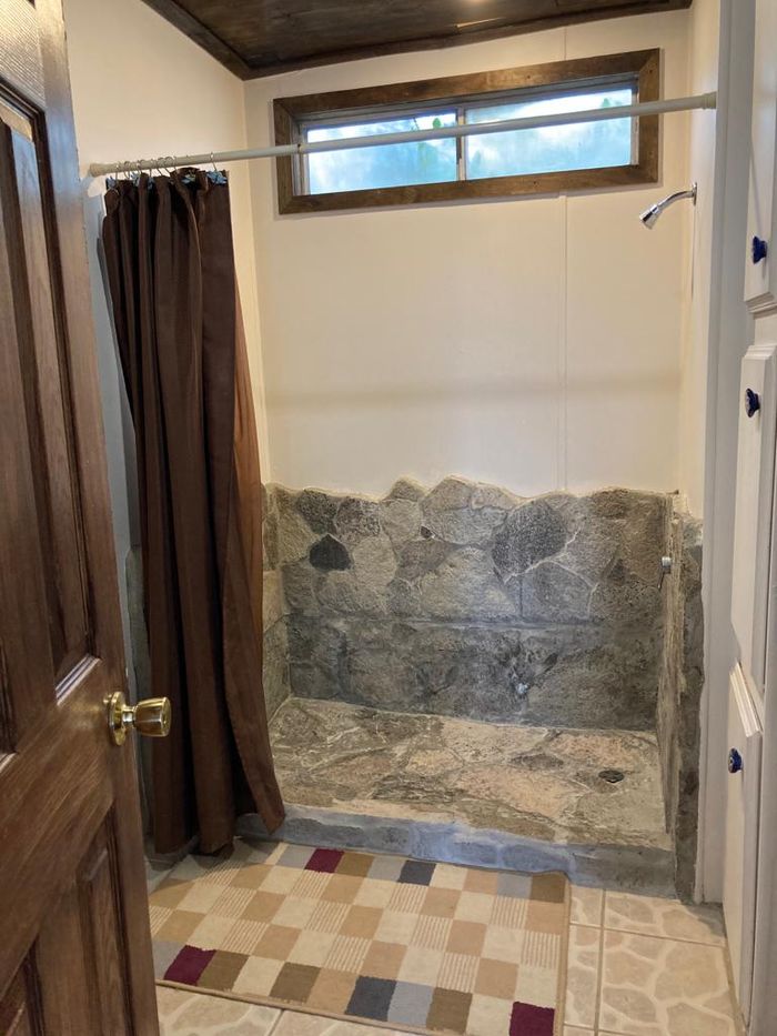 Baño pequeño con suelo de baldosas, zona de ducha con cortina y una pared decorativa de piedra debajo de una ventana.