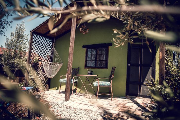 Casita de jardín verde con porche, silla hamaca y mesa exterior bajo un toldo frondoso.