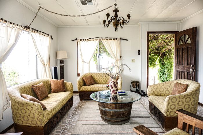 Luminoso salón con sofás de mimbre, sillones estampados, mesa de centro y puerta abierta que da a un jardín con vegetación.