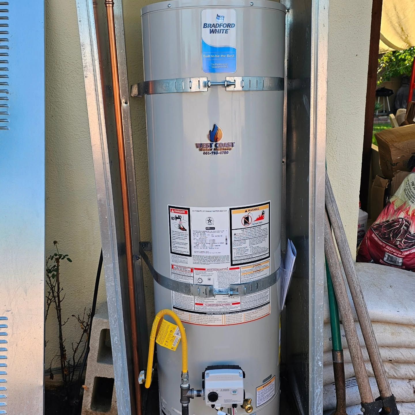 Silver water heater tank with warning labels and yellow hose in a narrow utility alcove