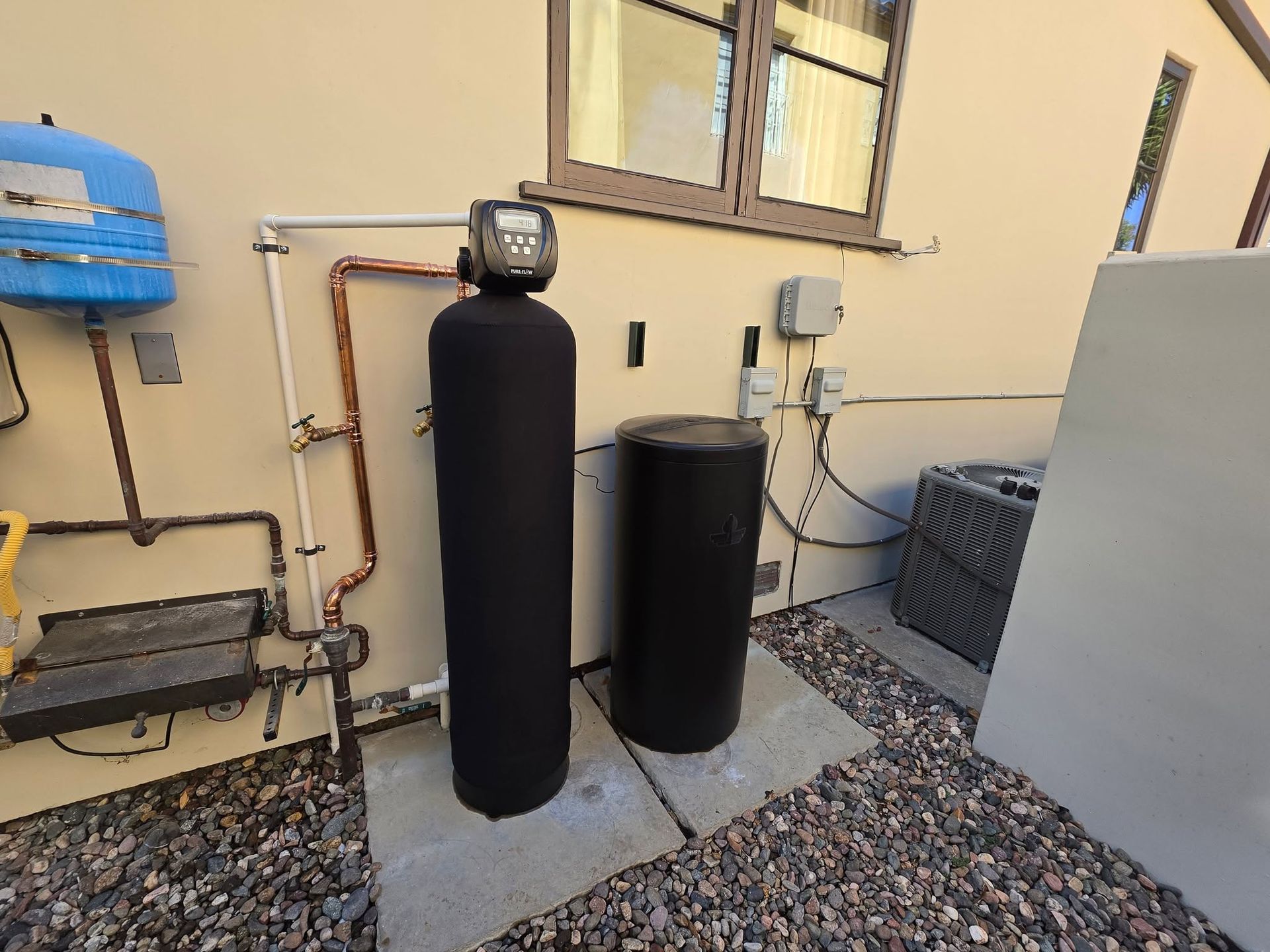Outdoor utility area with black water tanks, pipes, and a blue wall-mounted unit on gravel.